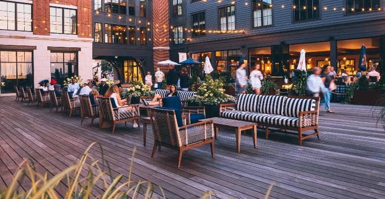Outdoor patio of a hotel in Newport, RI with people dining, featuring wooden furniture and striped cushions, surrounded by potted plants, at dusk.