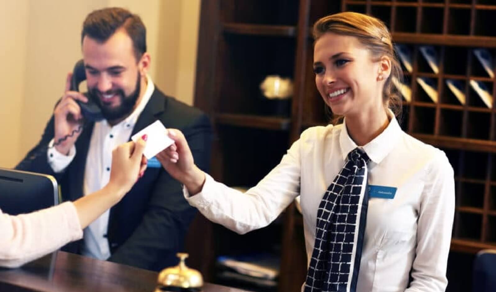 A female hotel receptionist at a Newport RI hotel hands a key card to a guest while a male colleague talks on a phone in the background.